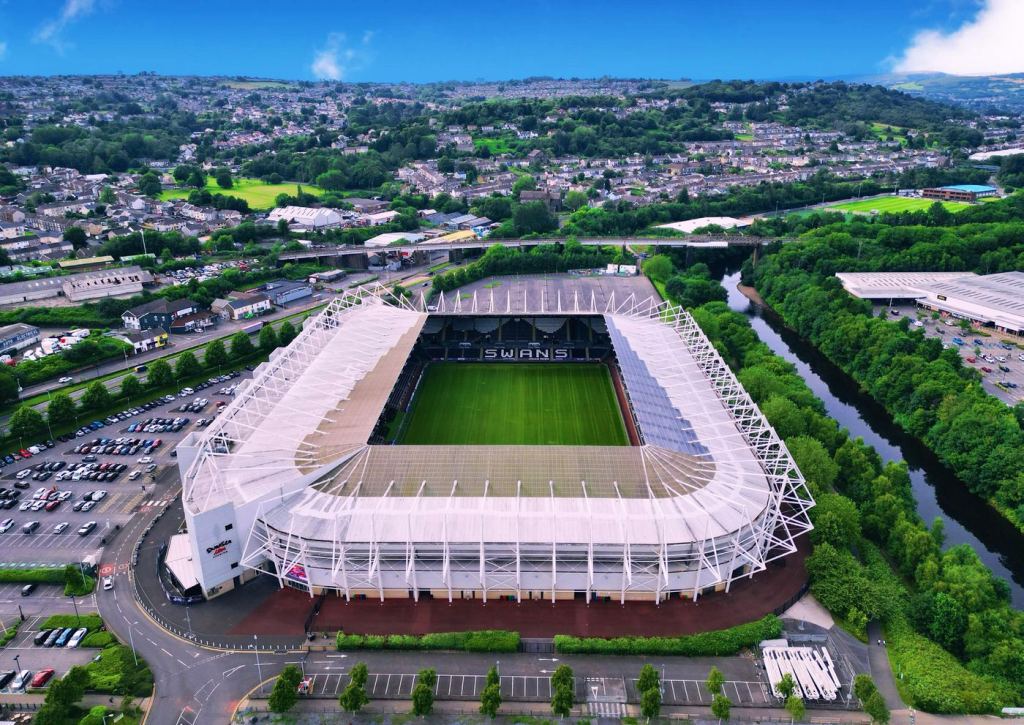 Aerial view of the Swansea.com Stadium with its white roof frame, car parks on one side and the River Tawe running alongside