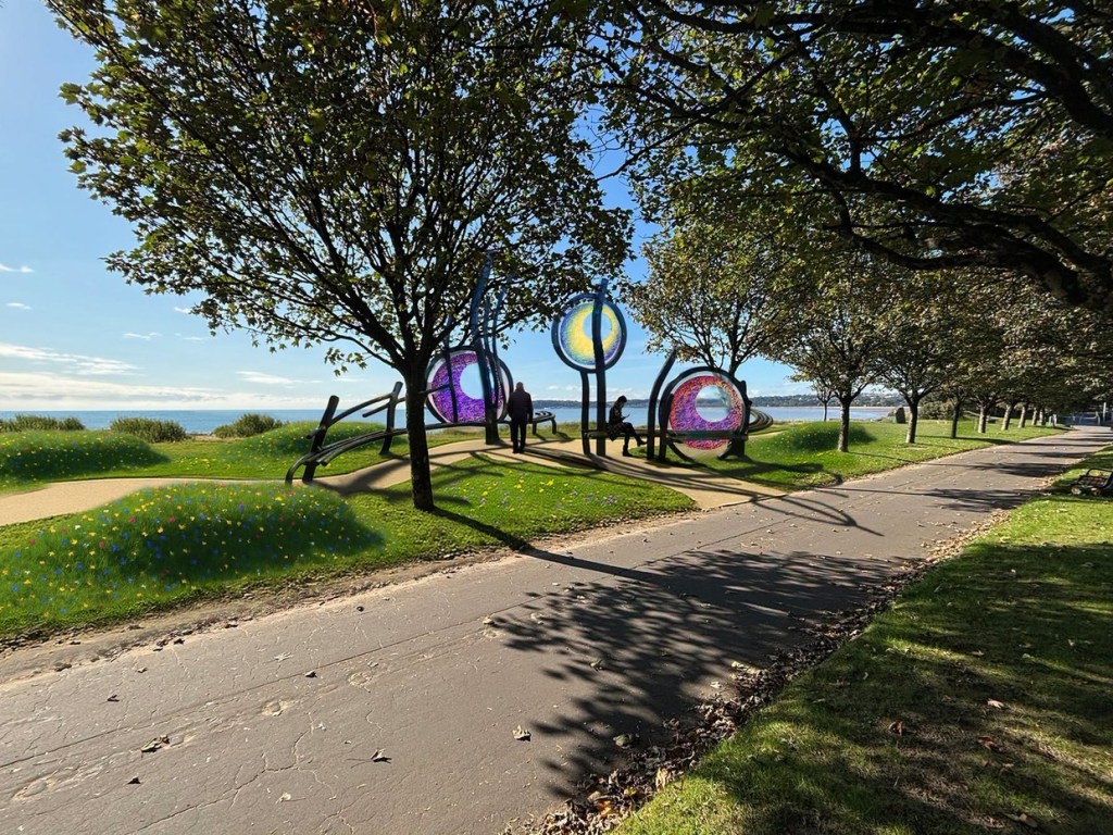 Artist's impression of three circular coloured glass sculptures on curved steel frames between trees on a grassy promenade, with wildflowers and Swansea Bay behind