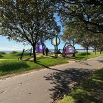 Artist's impression of three circular coloured glass sculptures on curved steel frames between trees on a grassy promenade, with wildflowers and Swansea Bay behind