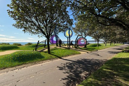 Artist's impression of three circular coloured glass sculptures on curved steel frames between trees on a grassy promenade, with wildflowers and Swansea Bay behind