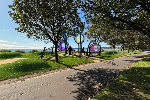 Artist's impression of three circular coloured glass sculptures on curved steel frames between trees on a grassy promenade, with wildflowers and Swansea Bay behind
