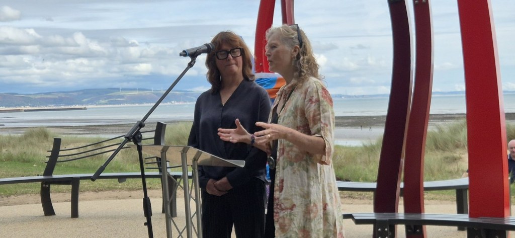 Two women speaking at a microphone beside the red steel memorial, with Swansea Bay behind them