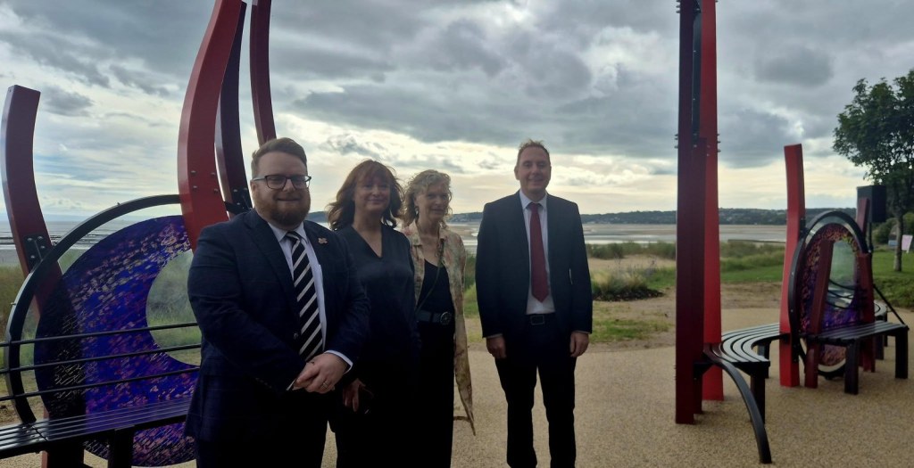 Two men and two women standing in front of the red steel and coloured glass Covid memorial on Swansea promenade