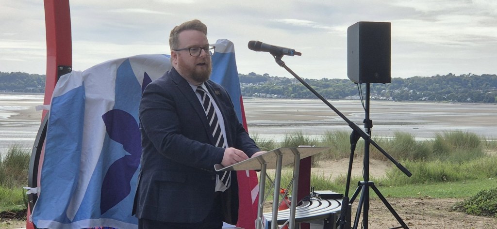 Cllr Elliott King speaking at a lectern in front of the covered memorial, with Swansea Bay behind
