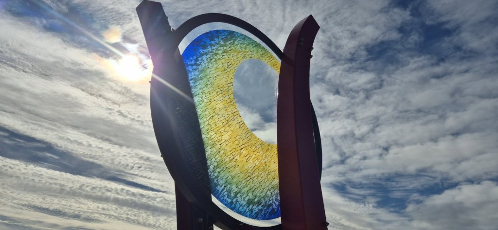 A circle of yellow and blue glass held between red steel uprights against a cloudy blue sky