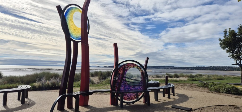 Curved benches around the red steel and glass memorial on the promenade, with Swansea Bay and the dunes behind