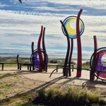 Red steel sculptures holding three circles of blue, yellow, purple and pink glass, surrounded by curved benches on the promenade with Swansea Bay behind