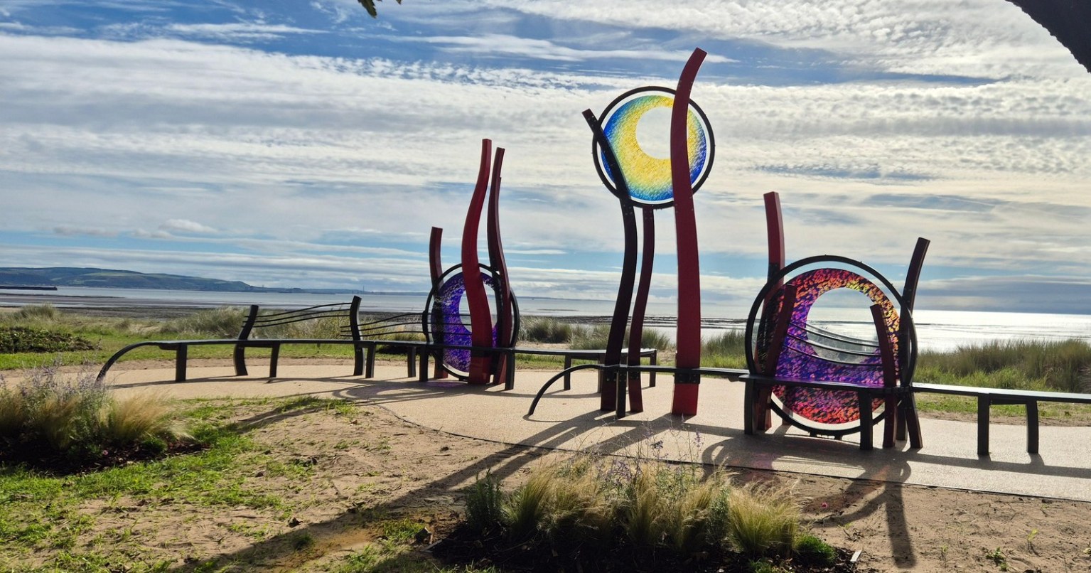 Red steel sculptures holding three circles of blue, yellow, purple and pink glass, surrounded by curved benches on the promenade with Swansea Bay behind