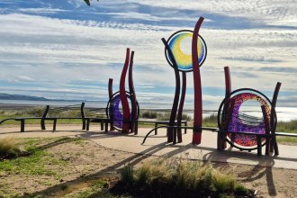 Red steel sculptures holding three circles of blue, yellow, purple and pink glass, surrounded by curved benches on the promenade with Swansea Bay behind
