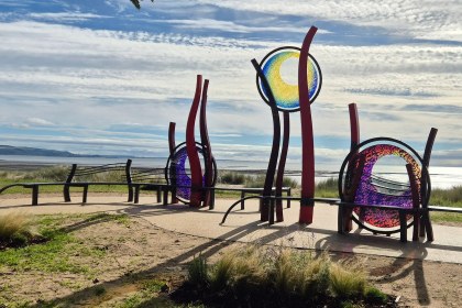 Red steel sculptures holding three circles of blue, yellow, purple and pink glass, surrounded by curved benches on the promenade with Swansea Bay behind