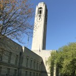 Swansea Guildhall's Portland stone clock tower against a clear blue sky, framed by spring trees, with the Welsh flag flying below