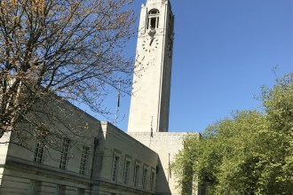 Swansea Guildhall's Portland stone clock tower against a clear blue sky, framed by spring trees, with the Welsh flag flying below