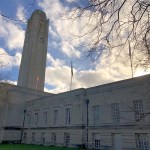 Swansea Guildhall's clock tower and stone frontage under a bright cloudy sky, with bare winter branches overhead and a Pride flag flying