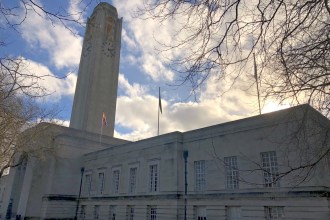 Swansea Guildhall's clock tower and stone frontage under a bright cloudy sky, with bare winter branches overhead and a Pride flag flying