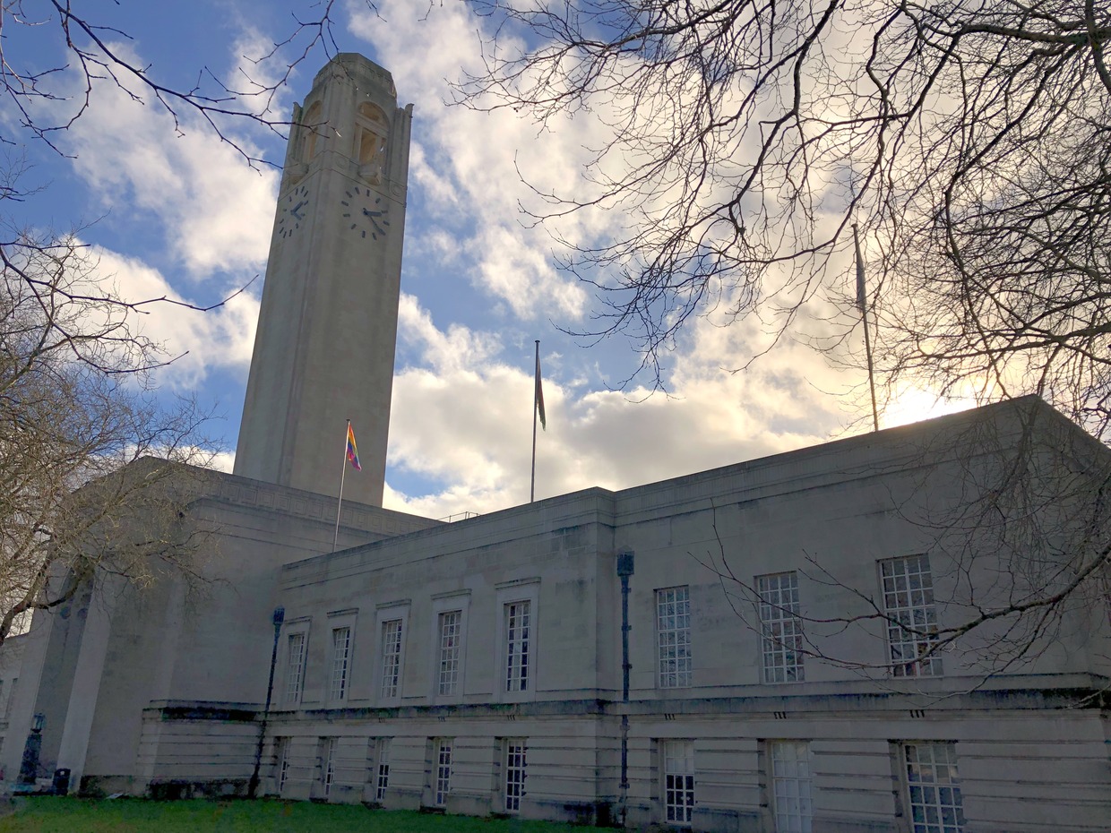 Swansea Guildhall's clock tower and stone frontage under a bright cloudy sky, with bare winter branches overhead and a Pride flag flying