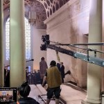 Film crew operating a camera crane on tracks inside the Guildhall's grand corridor in Swansea, with costumed actors beneath the honeycomb ceiling