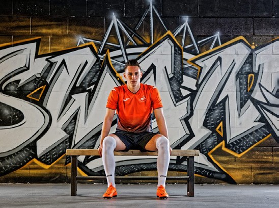 A player in an orange Swansea City kit sitting on a bench in front of a black and silver graffiti wall
