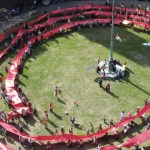 Aerial view of a long red banner coiled in concentric rings around a flagpole lawn outside Swansea Guildhall, held by marchers