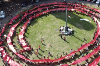 Aerial view of a long red banner coiled in concentric rings around a flagpole lawn outside Swansea Guildhall, held by marchers