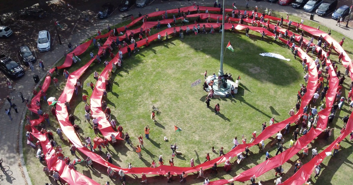 Aerial view of a long red banner coiled in concentric rings around a flagpole lawn outside Swansea Guildhall, held by marchers