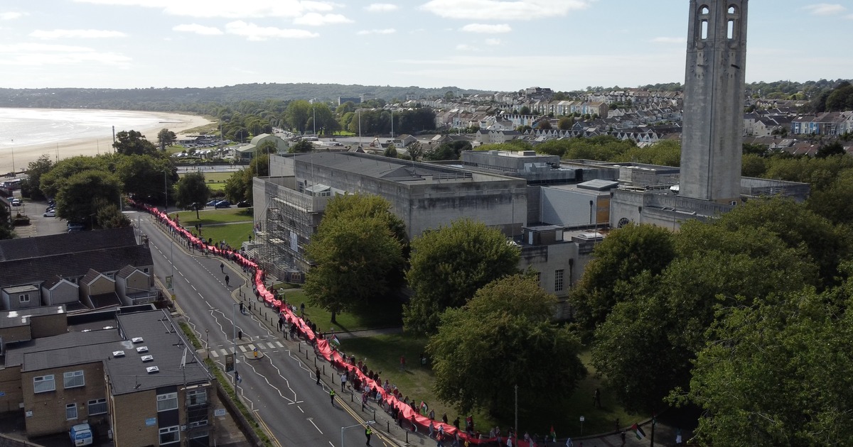 Aerial view of the red banner carried along the road past Swansea Guildhall and its clock tower, with Swansea Bay in the background