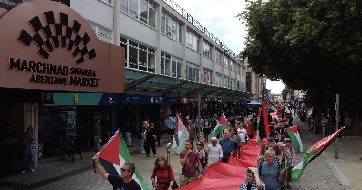 Marchers with Palestinian flags carrying a long red banner past the Swansea Market entrance on Oxford Street