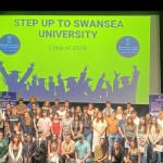 Around 70 young people pose on stage at the Taliesin Arts Centre in front of a screen reading Step Up to Swansea University, Class of 2026