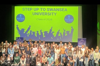 Around 70 young people pose on stage at the Taliesin Arts Centre in front of a screen reading Step Up to Swansea University, Class of 2026