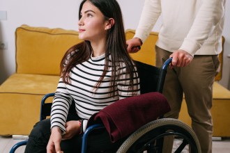 A young woman in a wheelchair at home, with a person standing behind pushing the chair