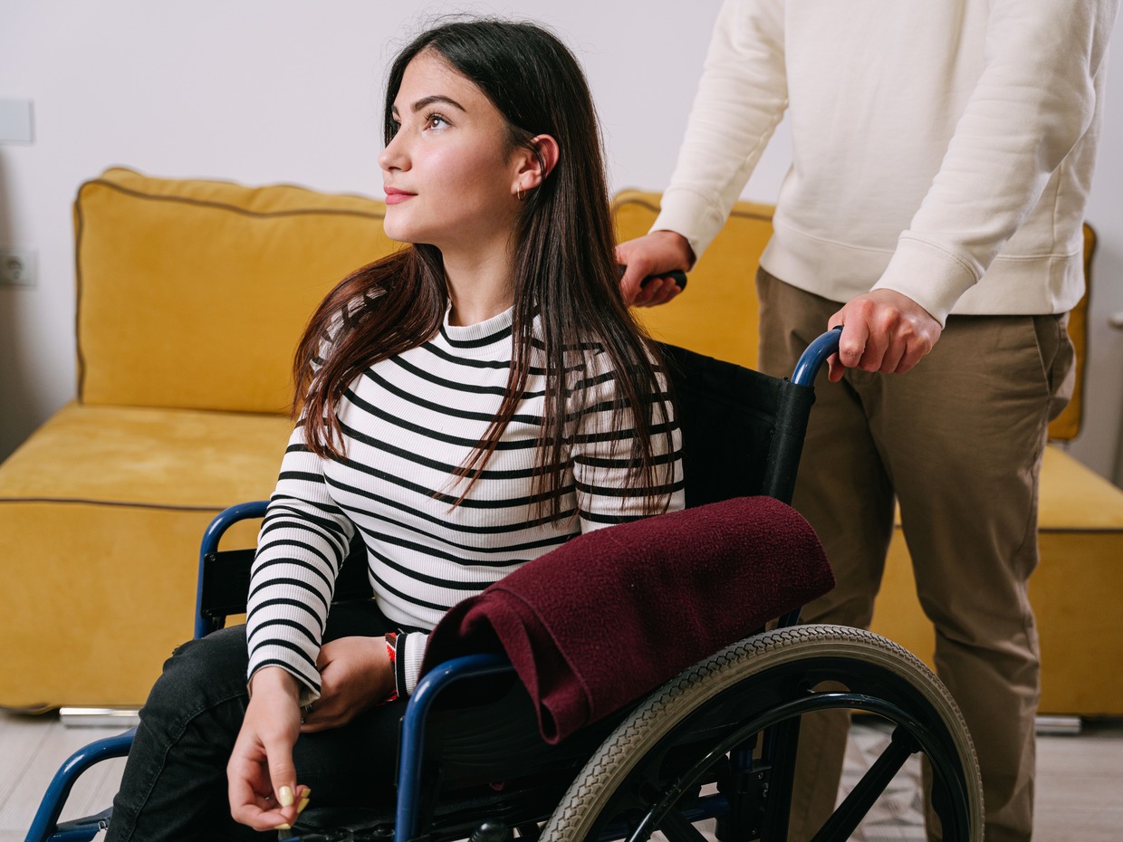 A young woman in a wheelchair at home, with a person standing behind pushing the chair