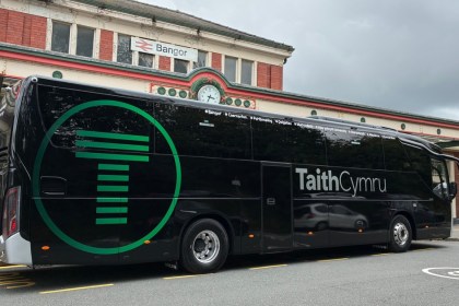 A black TaithCymru coach with a large green T logo parked outside Bangor railway station, the route's stops from Bangor to Carmarthen listed along its side