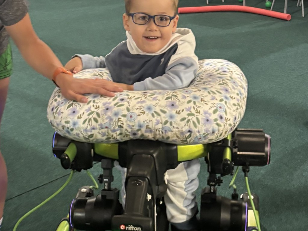 A three-year-old boy in glasses smiling with a hand to his mouth, standing in a green walking frame with glowing Trexo motor units at his hips and knees