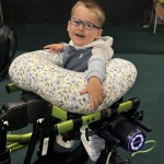 A smiling three-year-old boy in glasses standing in a green walking frame fitted with motorised Trexo leg units, resting on a floral cushion