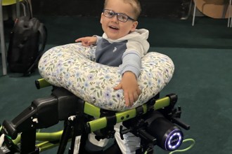 A smiling three-year-old boy in glasses standing in a green walking frame fitted with motorised Trexo leg units, resting on a floral cushion