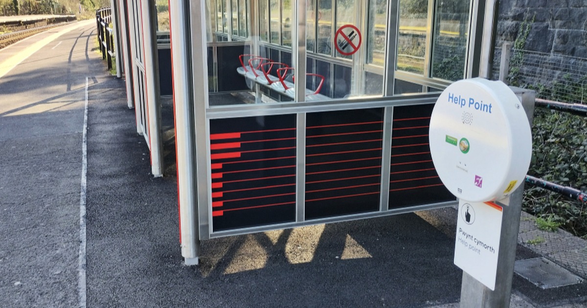 A white circular Transport for Wales help point unit mounted beside a station shelter on a platform