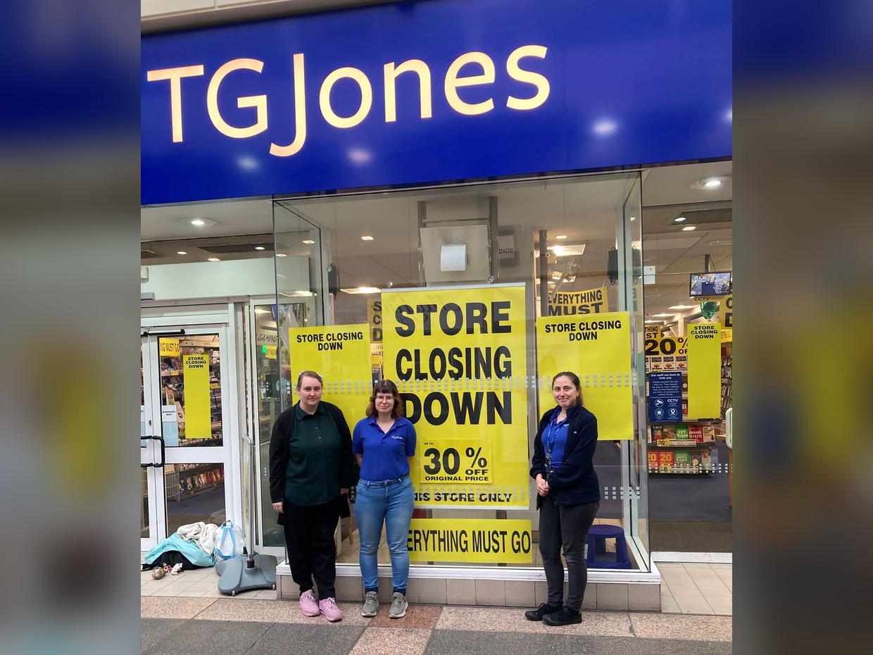 Three shop staff standing in front of the TGJones store in Bridgend's Rhiw Shopping Centre, with yellow 'store closing down, up to 30% off' posters filling the window behind them