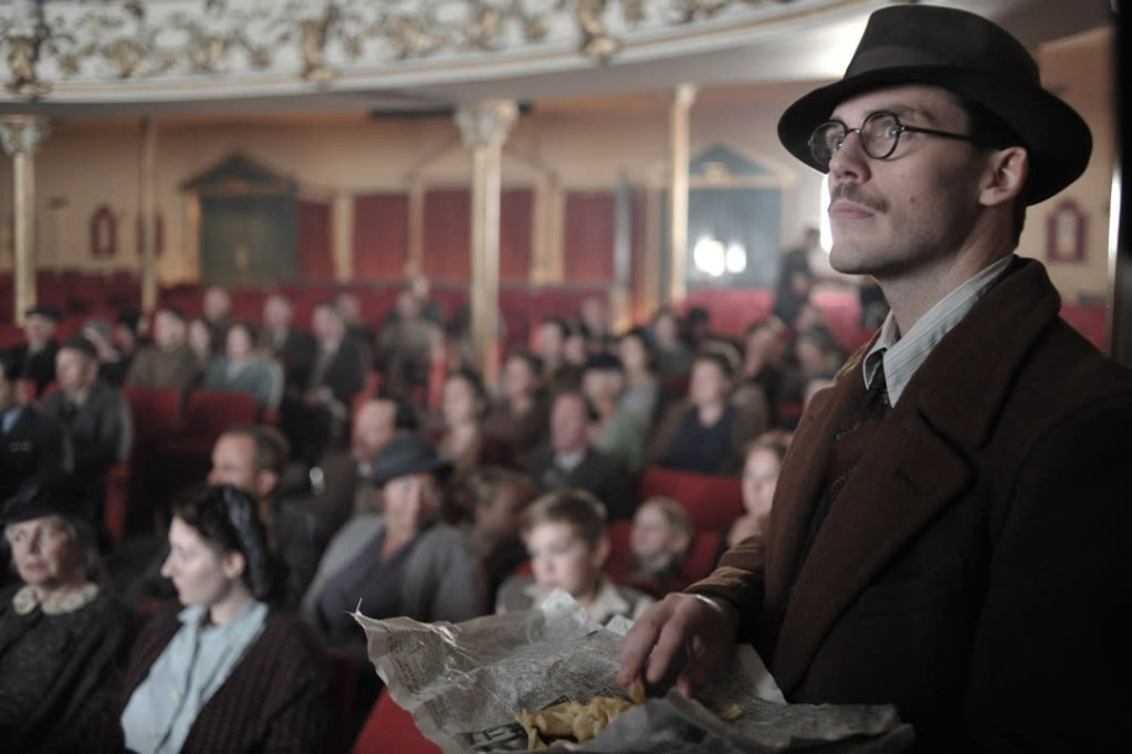 Sam Claflin in a trilby and glasses holding a newspaper of chips in the Swansea Grand Theatre auditorium, with a 1940s-costumed audience seated behind him