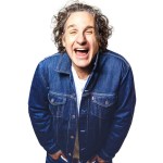 Comedian Tom Stade laughing, with curly grey hair and a dark denim jacket over a white T-shirt, against a white background