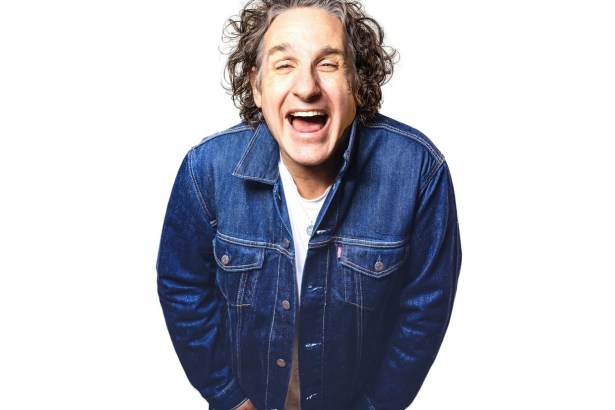 Comedian Tom Stade laughing, with curly grey hair and a dark denim jacket over a white T-shirt, against a white background