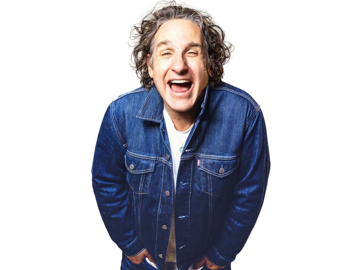 Comedian Tom Stade laughing, with curly grey hair and a dark denim jacket over a white T-shirt, against a white background