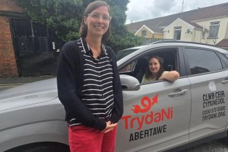 A woman standing beside a grey Hyundai Kona in TrydaNi Abertawe livery, with another woman at the wheel