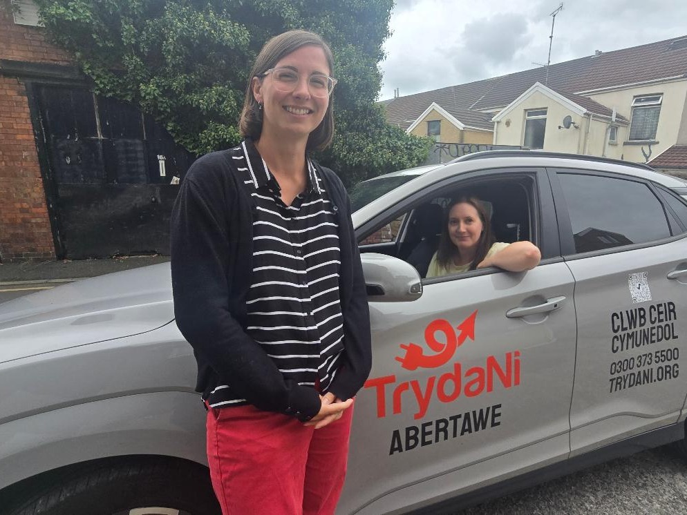 A woman standing beside a grey Hyundai Kona in TrydaNi Abertawe livery, with another woman at the wheel