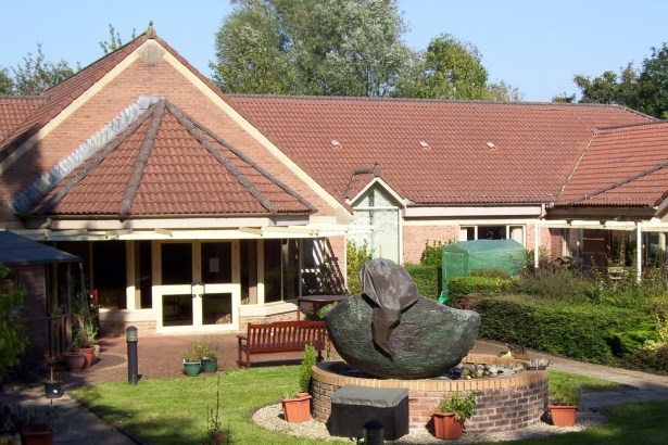 A single-storey red-brick hospice building with a tiled roof, with a large bronze sculpture on a circular brick plinth and a wooden bench in the garden in front