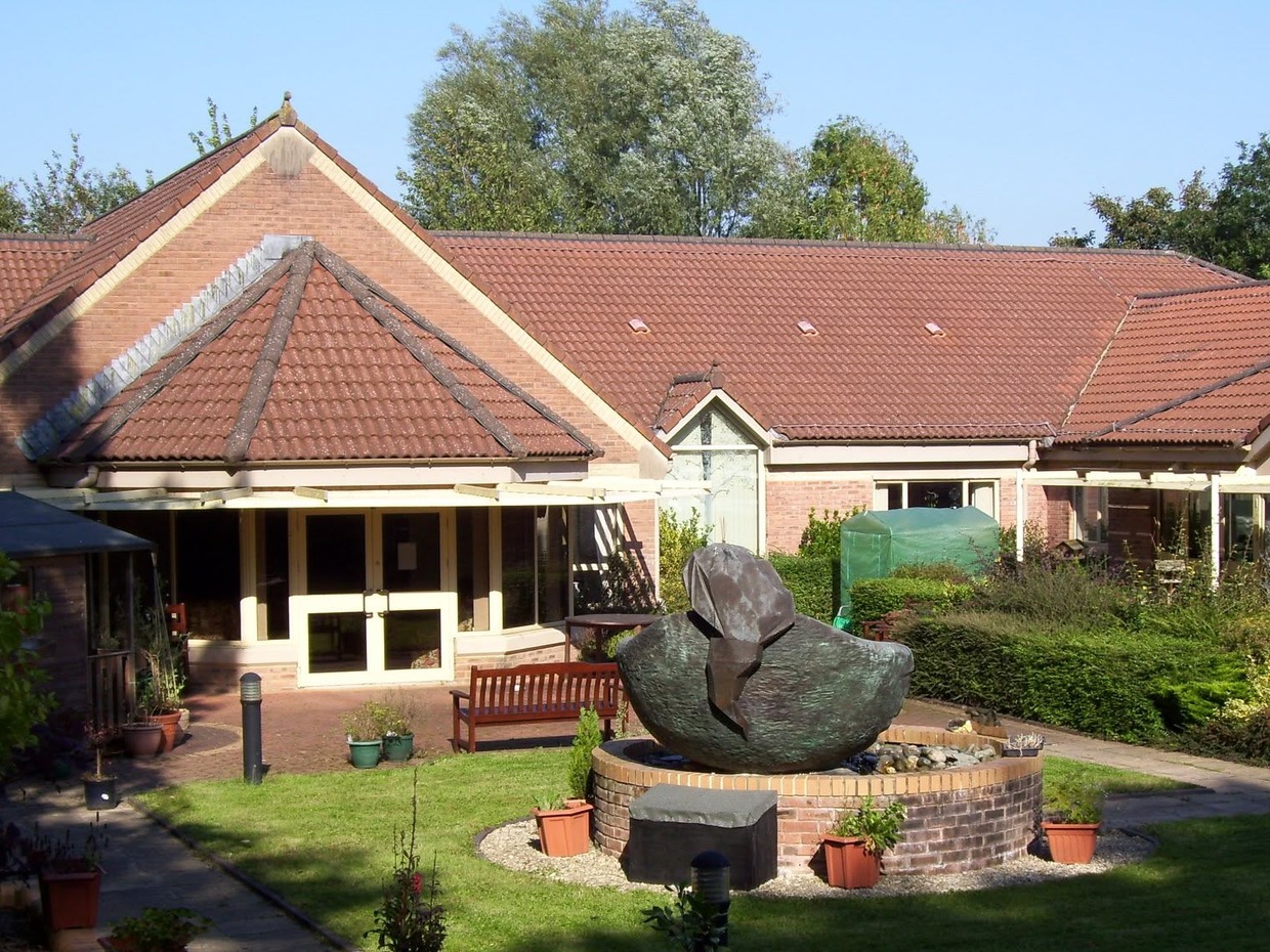 A single-storey red-brick hospice building with a tiled roof, with a large bronze sculpture on a circular brick plinth and a wooden bench in the garden in front
