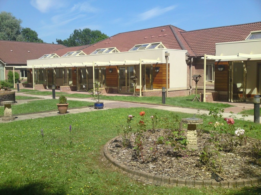 A low red-roofed building with a row of glazed doors under a cream pergola, opening onto a lawn with rose beds, bird tables and a sundial