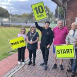 Five people beside a bowling green in Llandybie holding yellow race signs reading Race HQ, 10 KMS and Start, one wearing a chain of office, with tennis courts behind