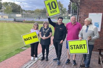 Five people beside a bowling green in Llandybie holding yellow race signs reading Race HQ, 10 KMS and Start, one wearing a chain of office, with tennis courts behind