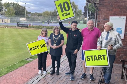 Five people beside a bowling green in Llandybie holding yellow race signs reading Race HQ, 10 KMS and Start, one wearing a chain of office, with tennis courts behind