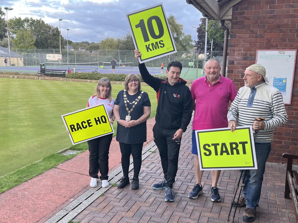 Five people beside a bowling green in Llandybie holding yellow race signs reading Race HQ, 10 KMS and Start, one wearing a chain of office, with tennis courts behind