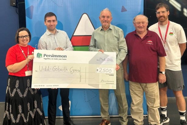 Five people standing in front of an Urdd banner holding a large Persimmon cheque made out to Urdd Gobaith Cymru for £2,500